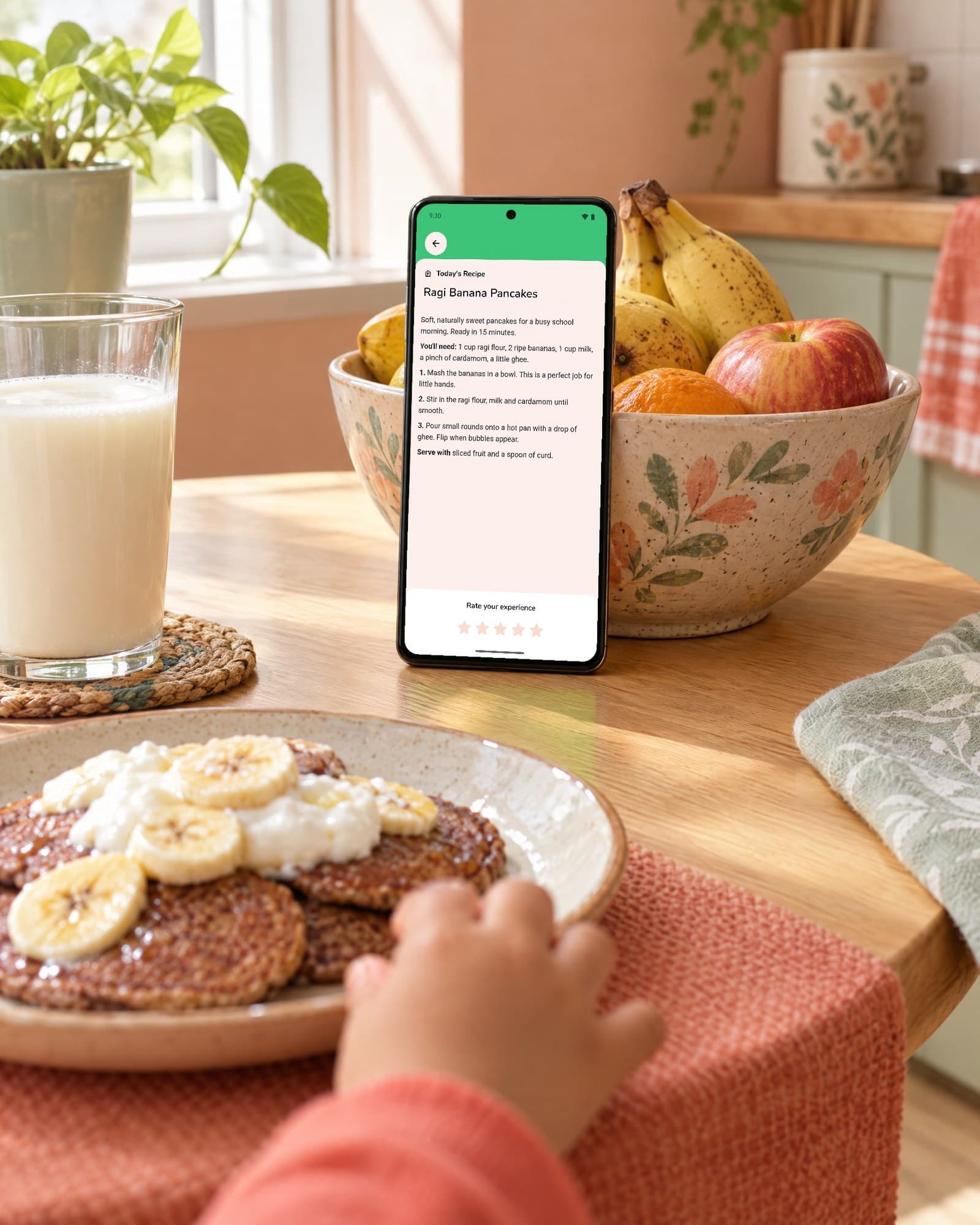 Morning at a sunny breakfast table: a phone stands against a bowl of bananas and apples, showing Today's Recipe, Ragi Banana Pancakes. In front, a plate of the pancakes with banana and curd, a glass of milk, and a small hand reaching for the plate