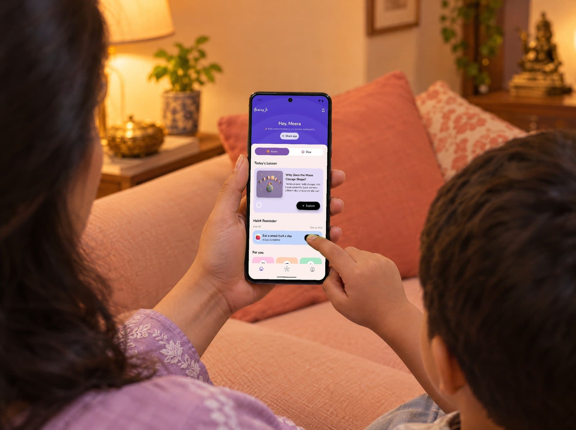 Evening on a peach sofa: a mother in a lavender kurta holds a phone while her son points at the screen. On it, Brainy Junior's home: Hey, Meera, the children's tabs with a lion cub for Aarav and a bunny for Diya, today's lesson, Why Does the Moon Change Shape?, and his habit, Eat a small fruit a day.