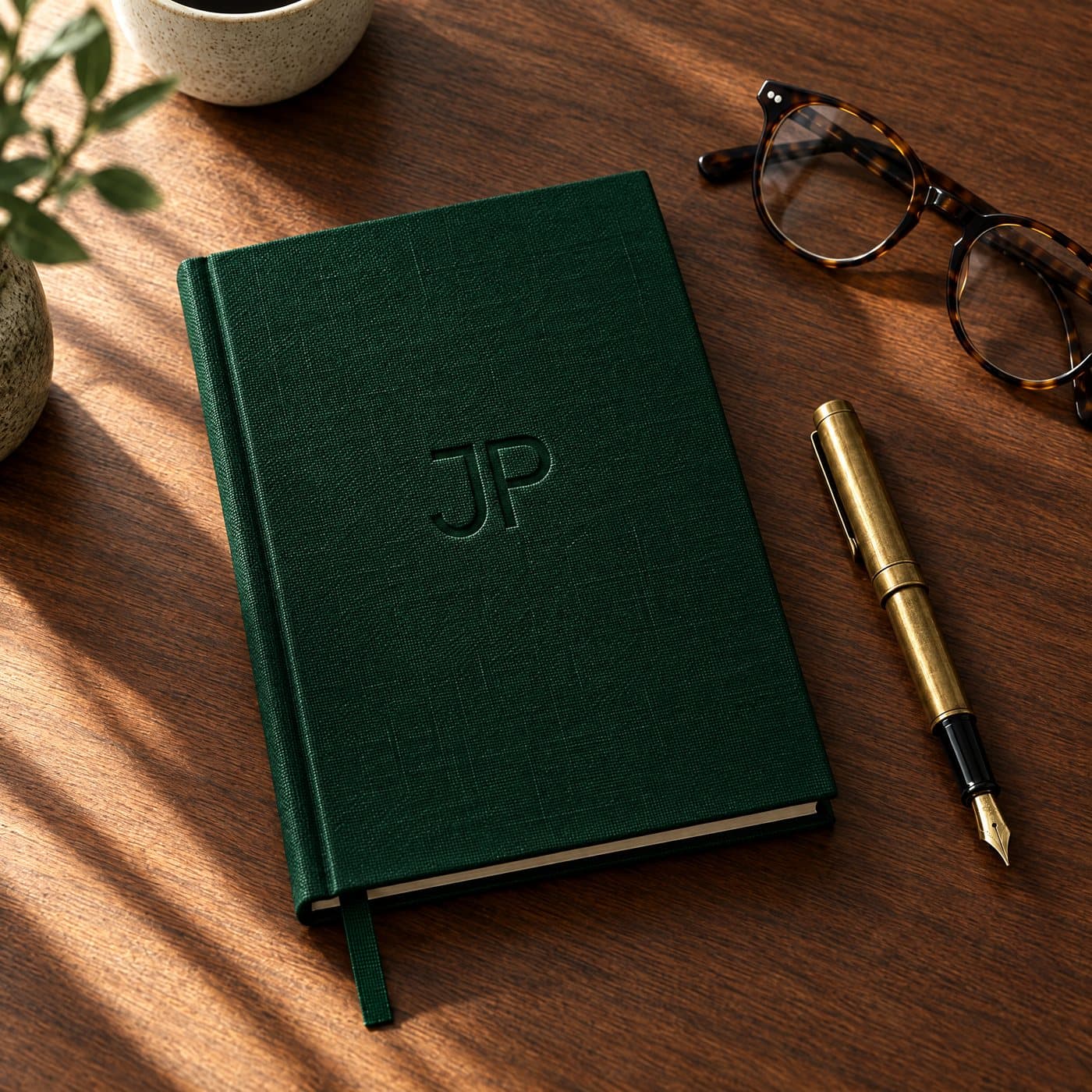 A deep green linen notebook with the JP monogram debossed, a brass pen and reading glasses on walnut